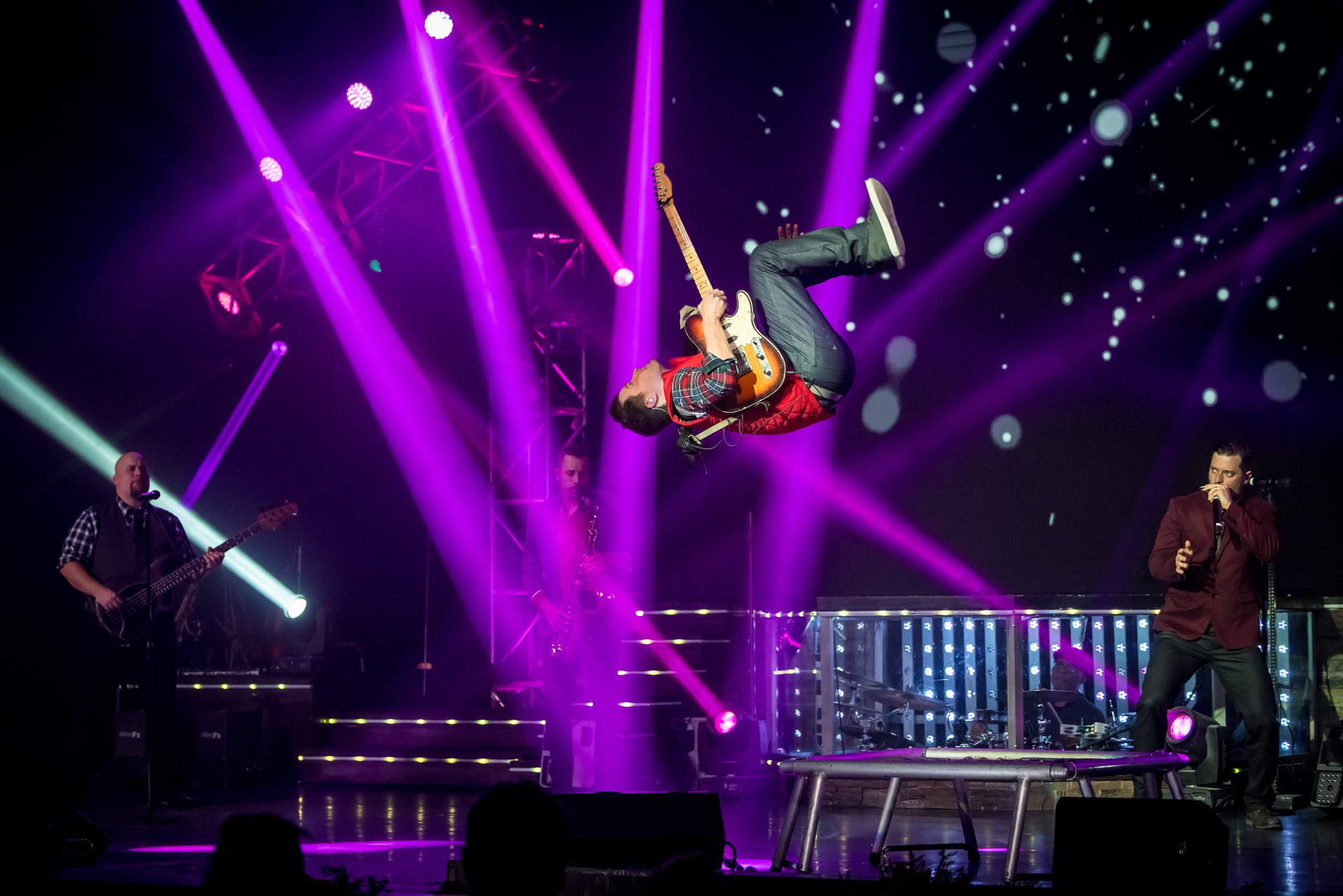A musician performs a backflip on stage while playing an electric guitar, surrounded by bright purple spotlights.
