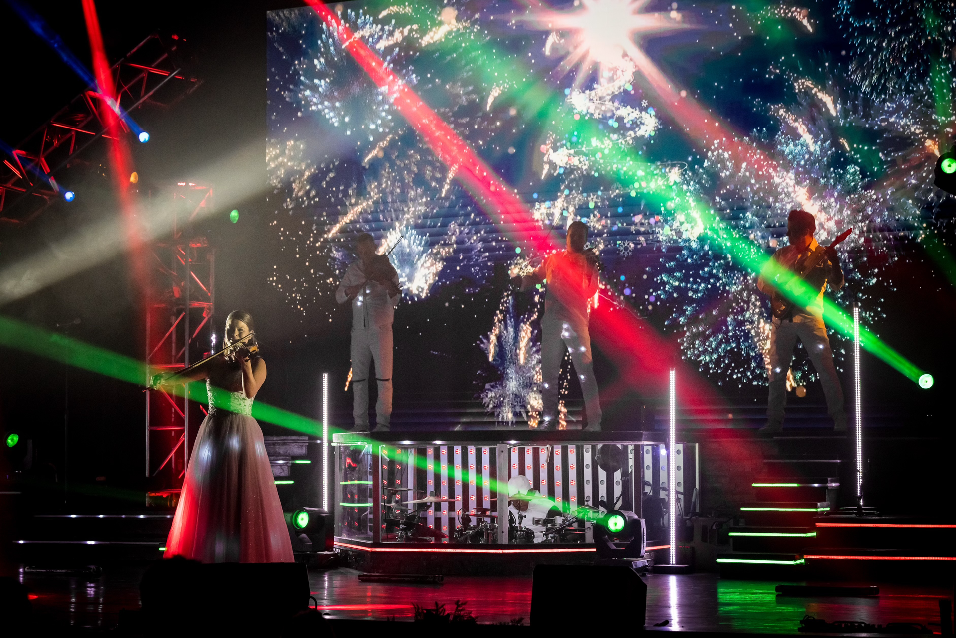 A performer in a long dress sings on a stage illuminated by bright green and red laser lights with a large video backdrop.