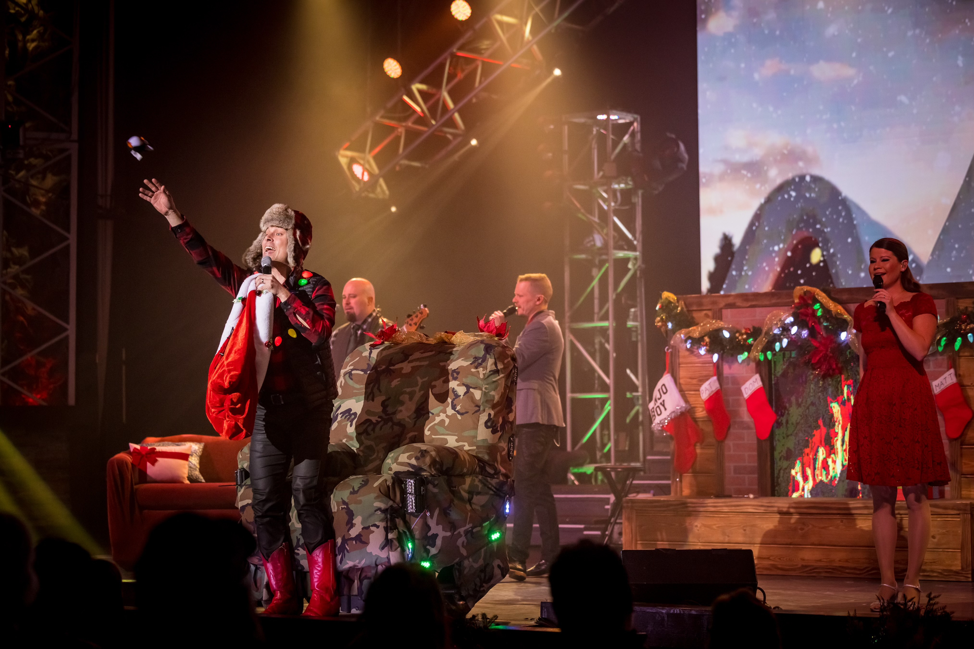 Performers on a stage with a fireplace backdrop and festive decor, one gesturing while holding a red stocking.