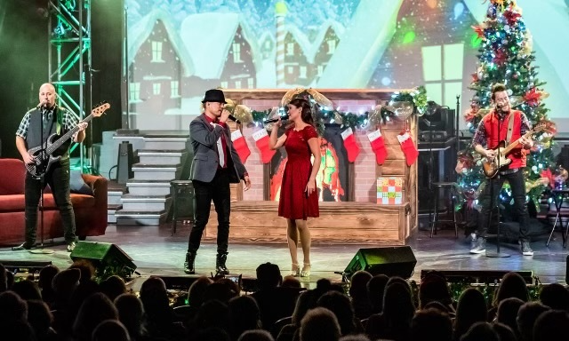 A band performs on a holiday-themed stage with a fireplace, stockings, and a Christmas tree, facing an audience.