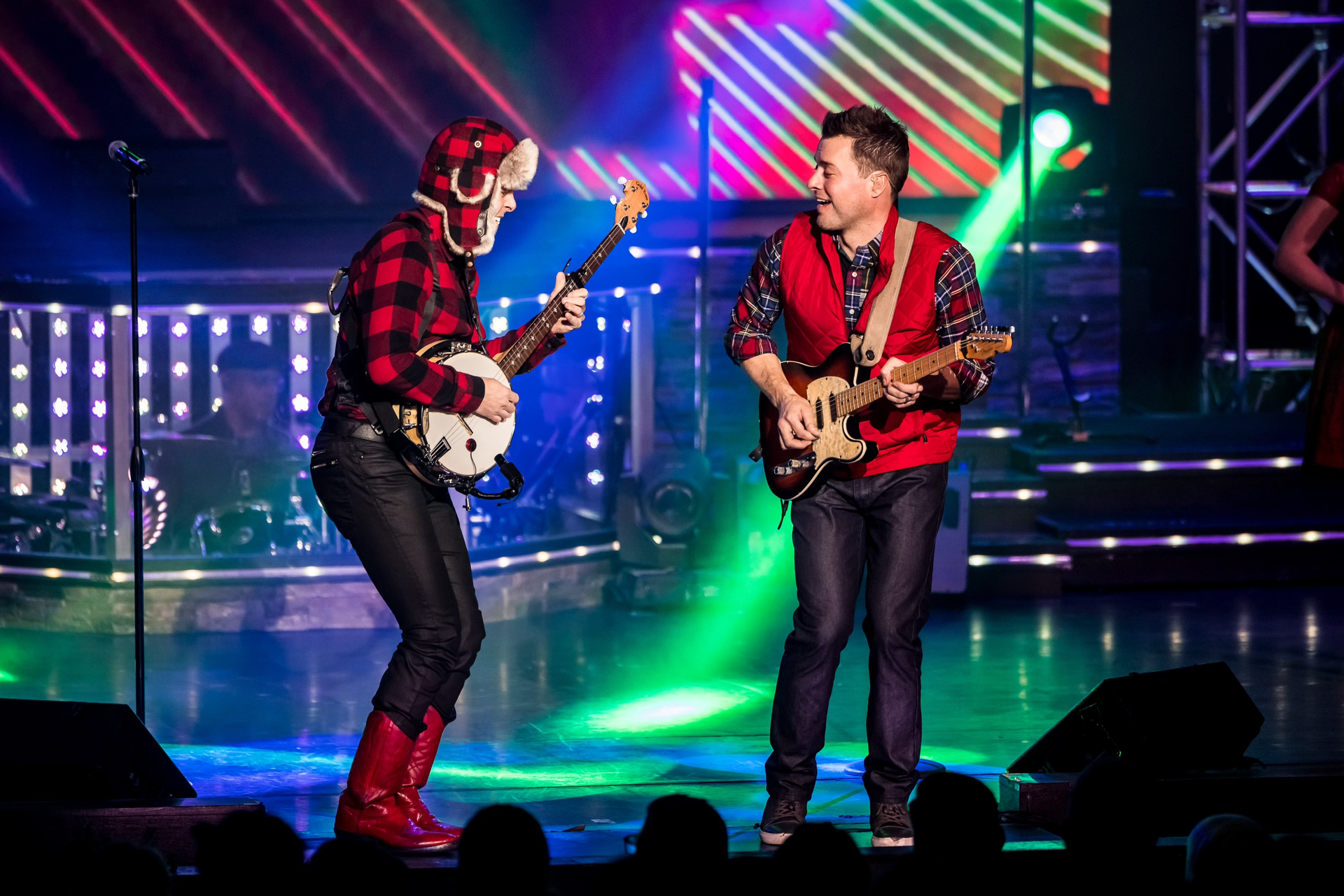 Two musicians perform on stage; one plays a banjo in a flannel outfit, the other plays an electric guitar in a red vest.