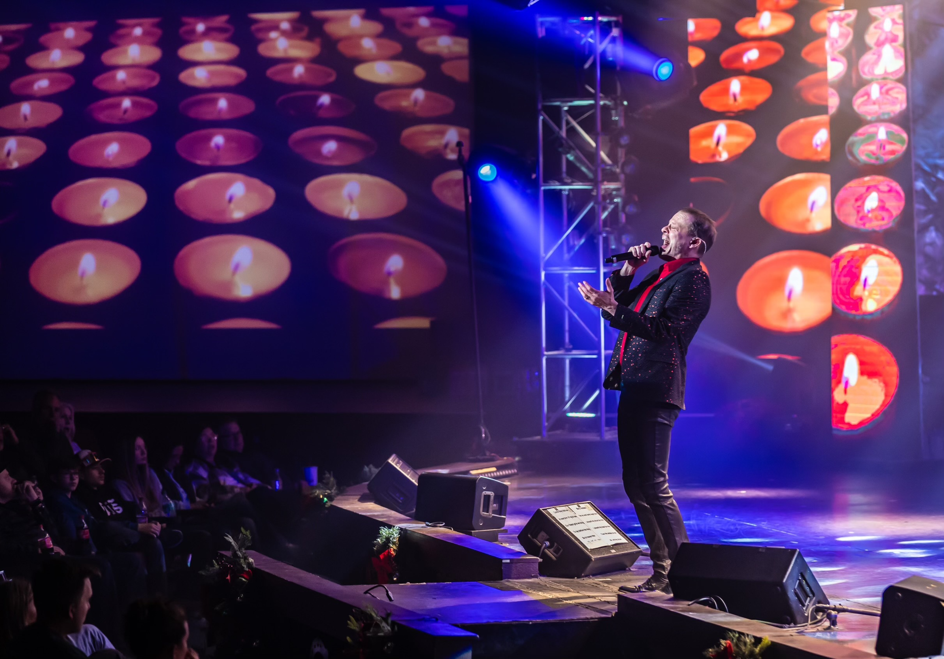 A performer sings on a stage illuminated by blue spotlights, with a large screen displaying many glowing candles behind.