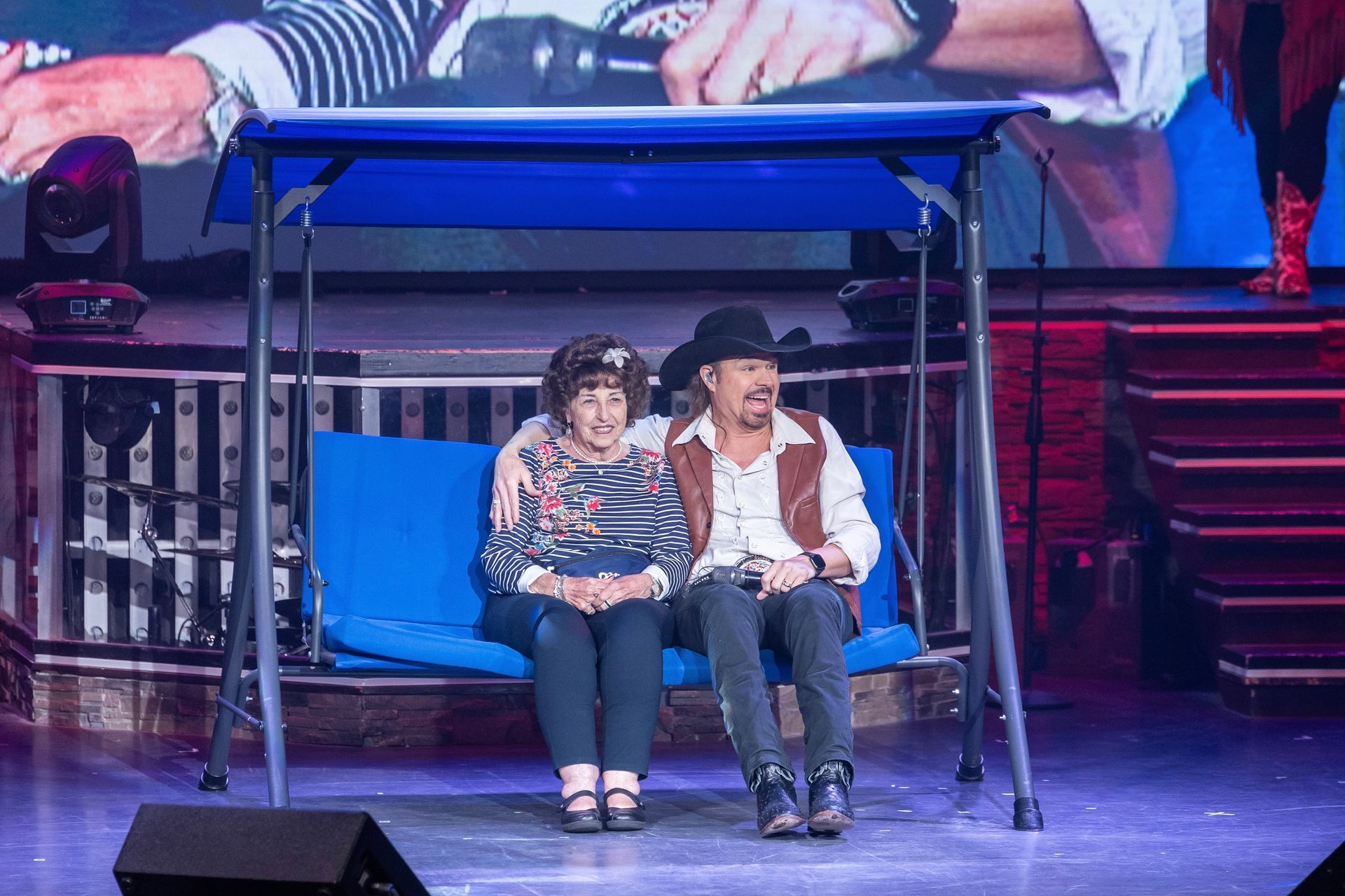 A woman and a man in a cowboy hat sit together on a blue porch swing on a stage with a large screen in the background.