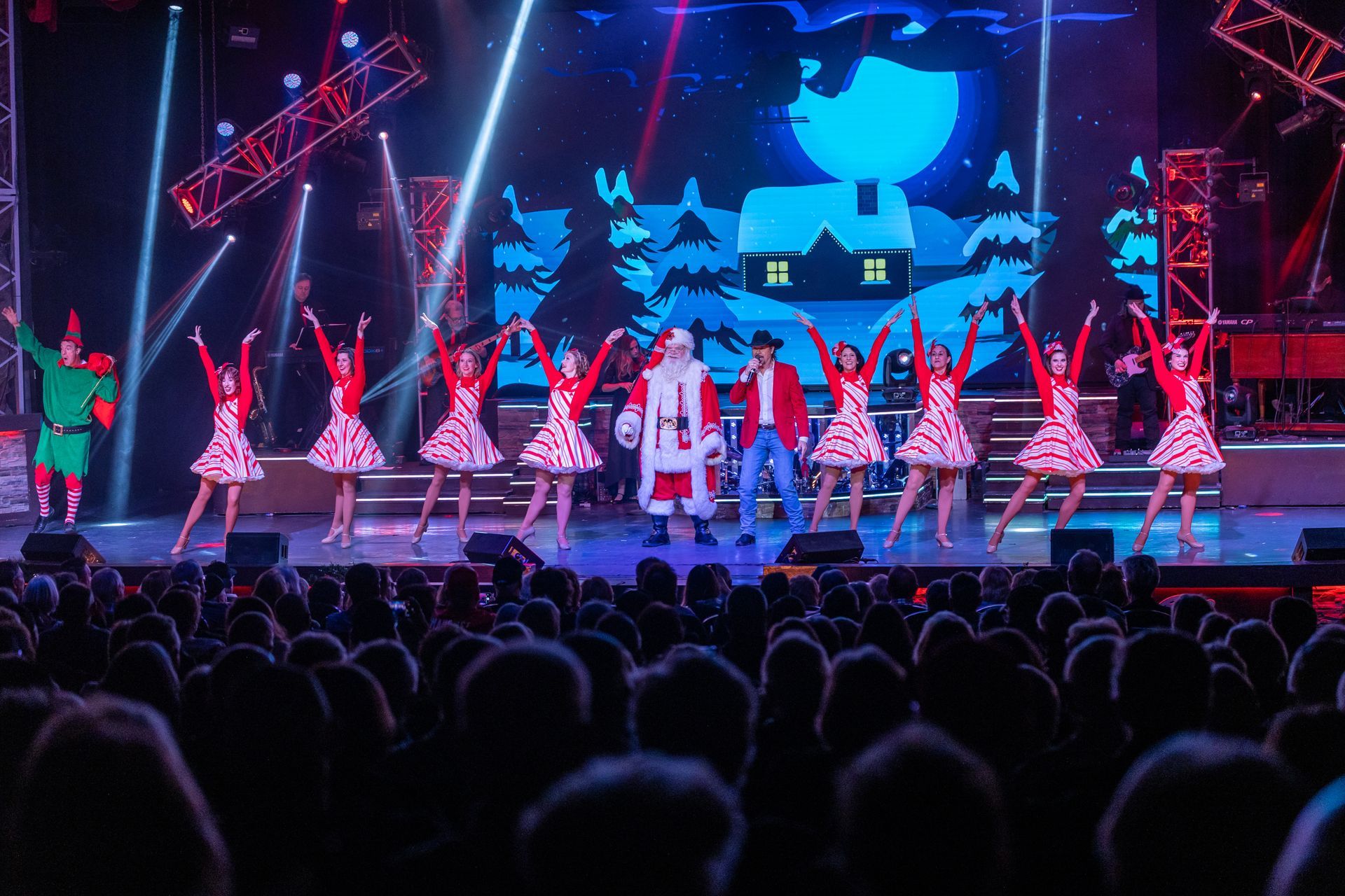 Performers in festive outfits, including Santa, dance on a stage before a large audience under bright spotlights.