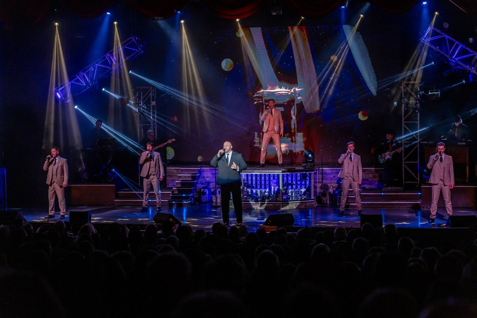 Five singers in light-colored suits perform on a stage with bright spotlights under a dark, starry backdrop.