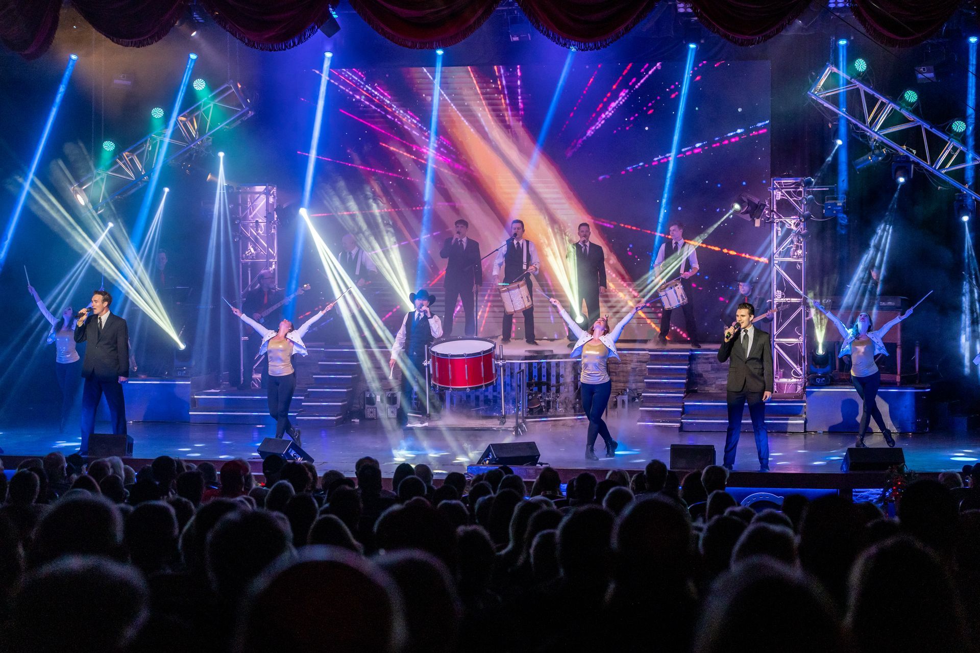 A band and dancers perform on a brightly lit, neon-blue stage before a large audience in a theater.