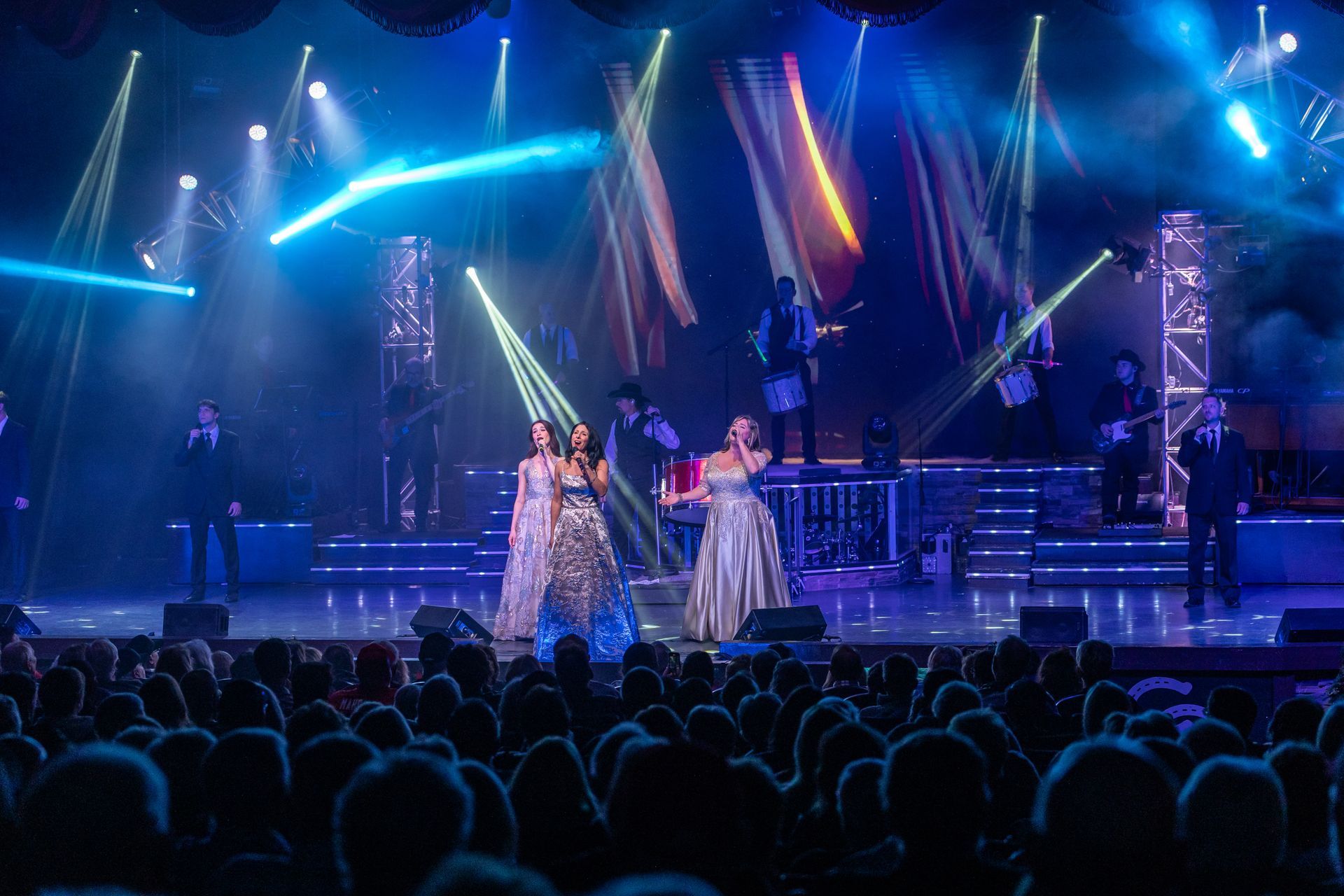Three performers in elegant gowns stand on a stage with bright blue spotlights, singing to a large audience.