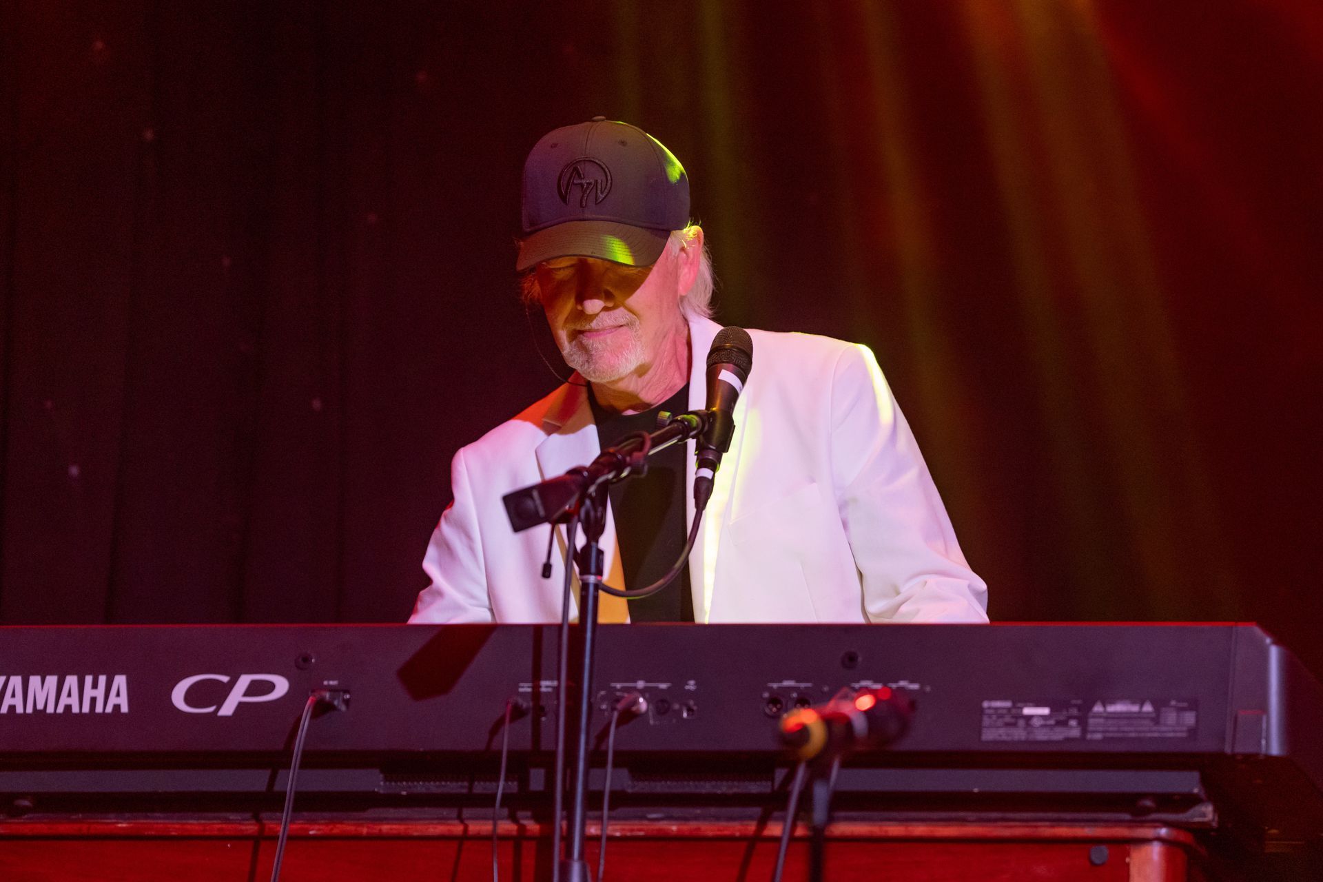 A person wearing a cap and white blazer plays a Yamaha CP keyboard on a dimly lit stage.