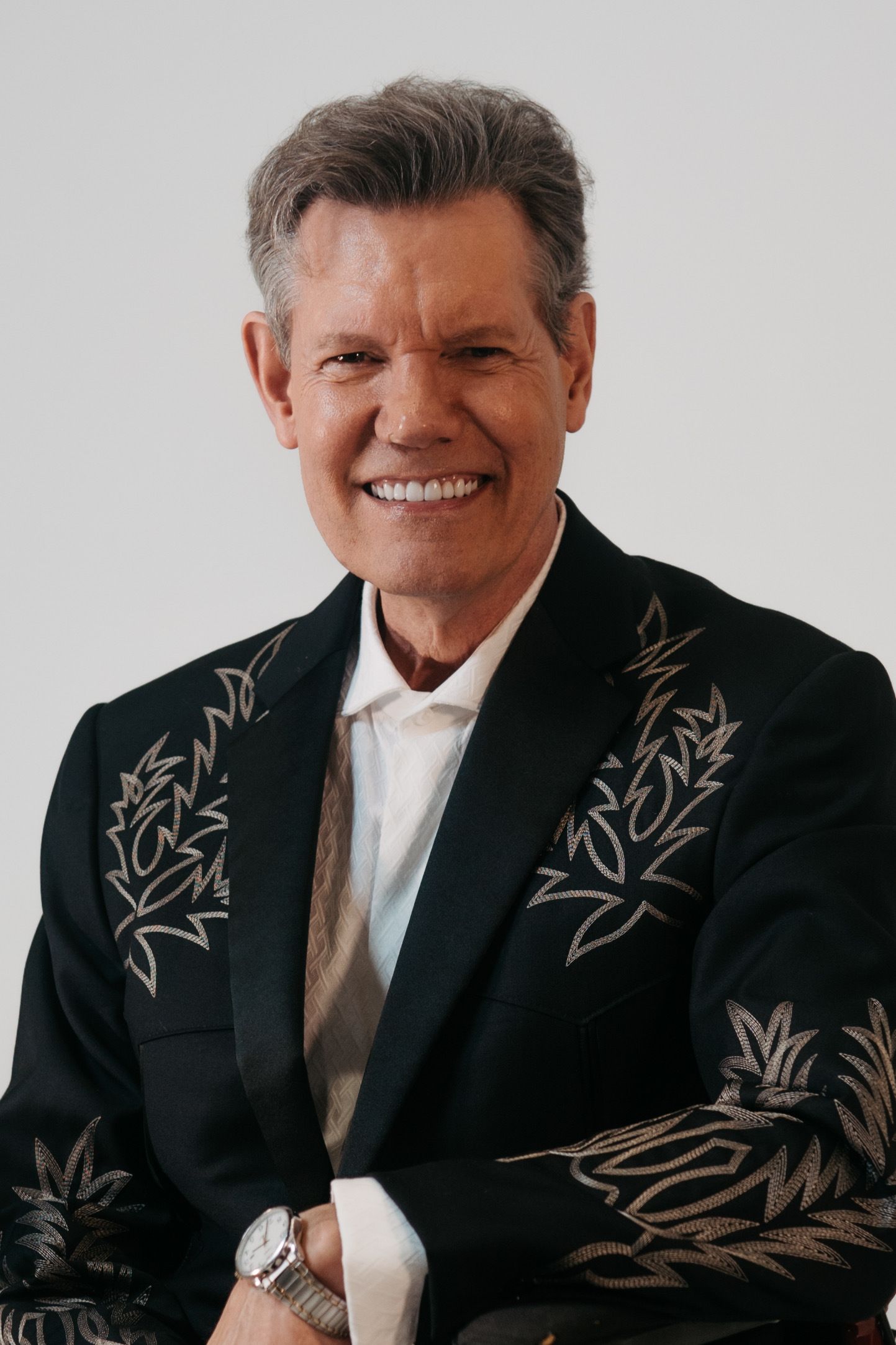 A smiling person in a black, embroidered blazer with a white collared shirt, set against a plain light background.