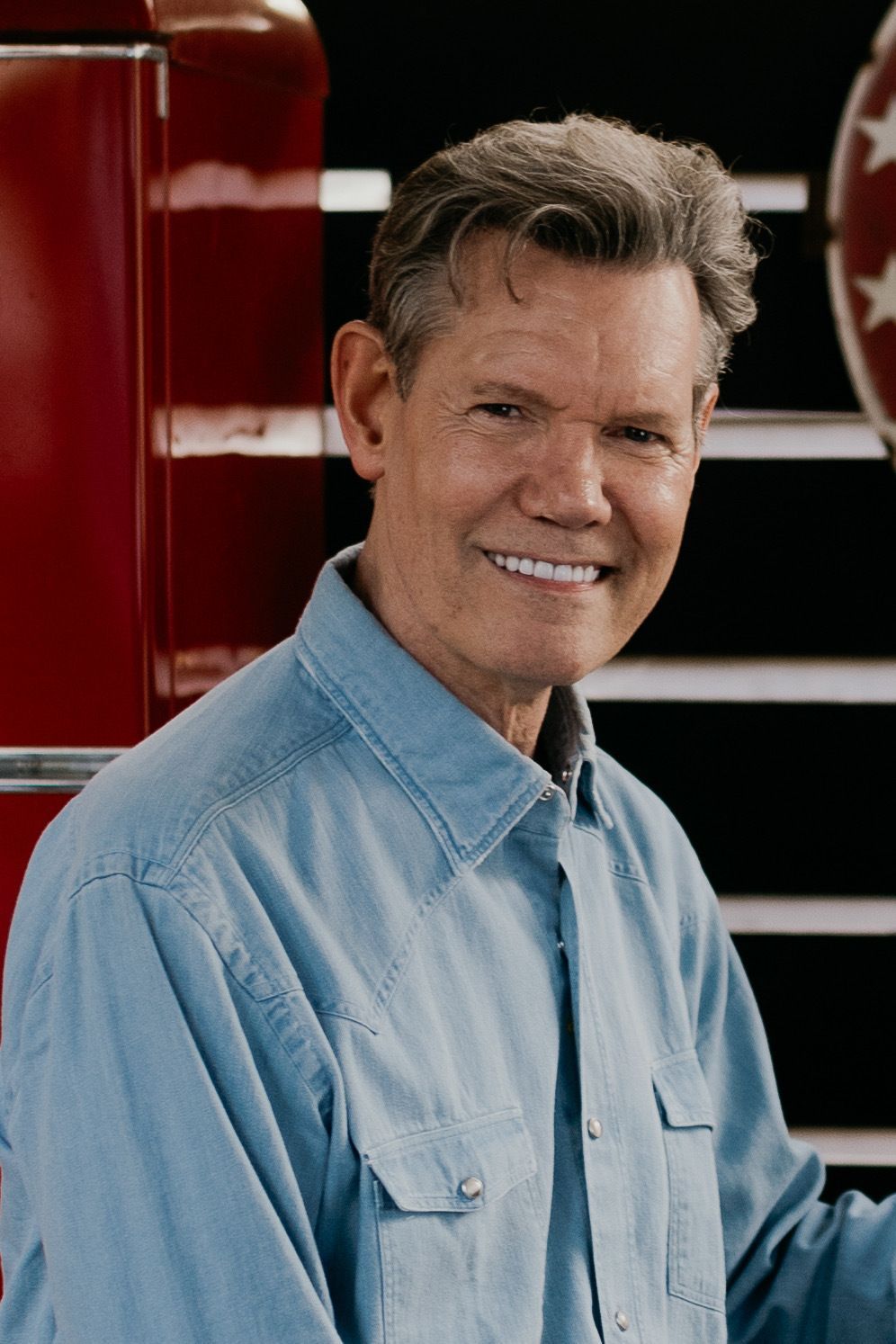 A smiling person in a blue denim button-down shirt standing in a workshop with a red cabinet in the background.