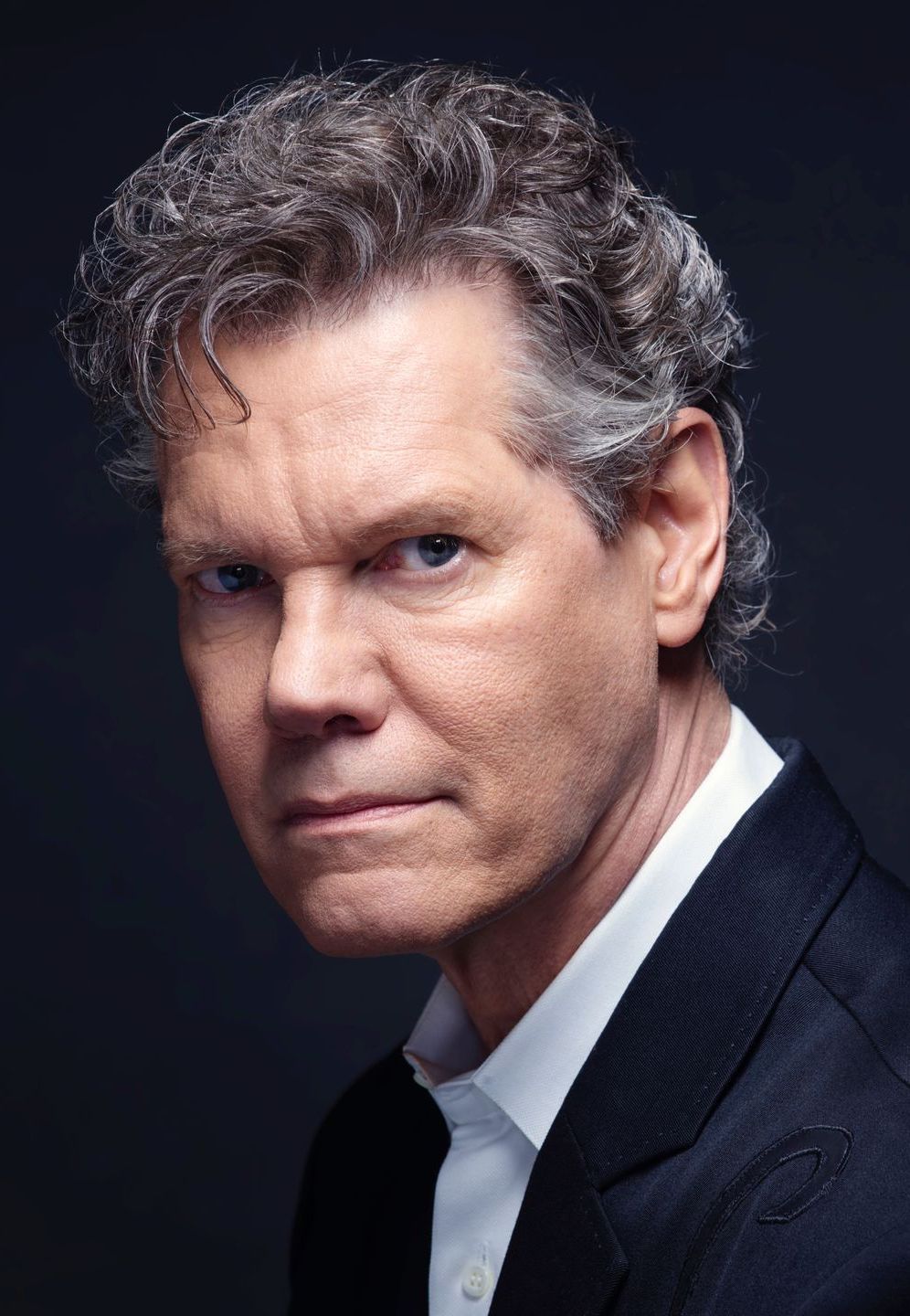 A person with wavy hair, wearing a white collared shirt and dark blazer, looks directly ahead against a black background.