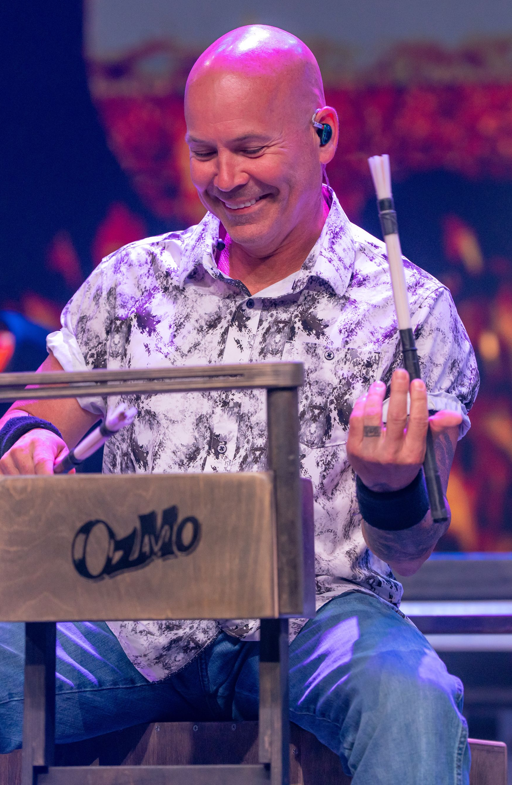 A smiling musician in a patterned shirt plays a wooden percussion instrument labeled 