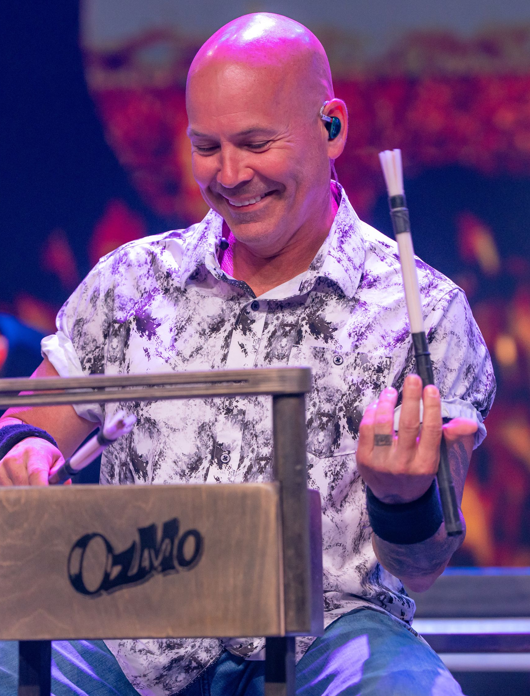 A smiling performer holds drum brushes while playing a wooden percussion instrument labeled