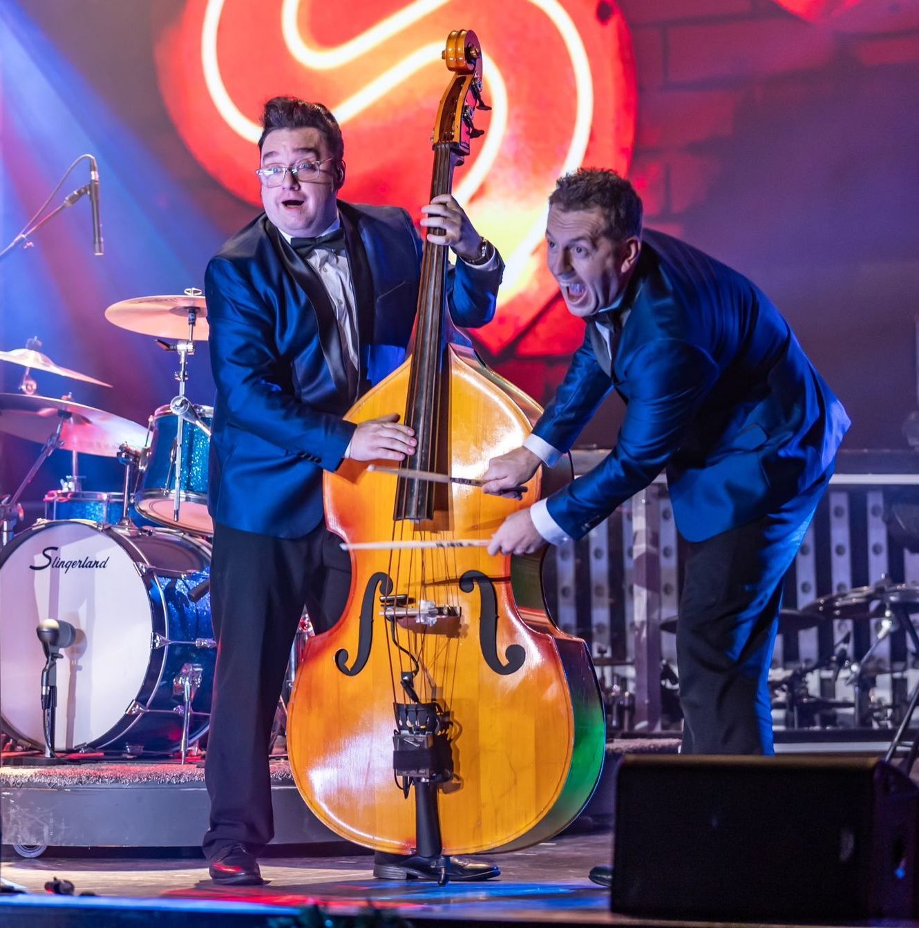 Two musicians in blue suits perform on stage, playing a double bass together with drumsticks in a concert setting.