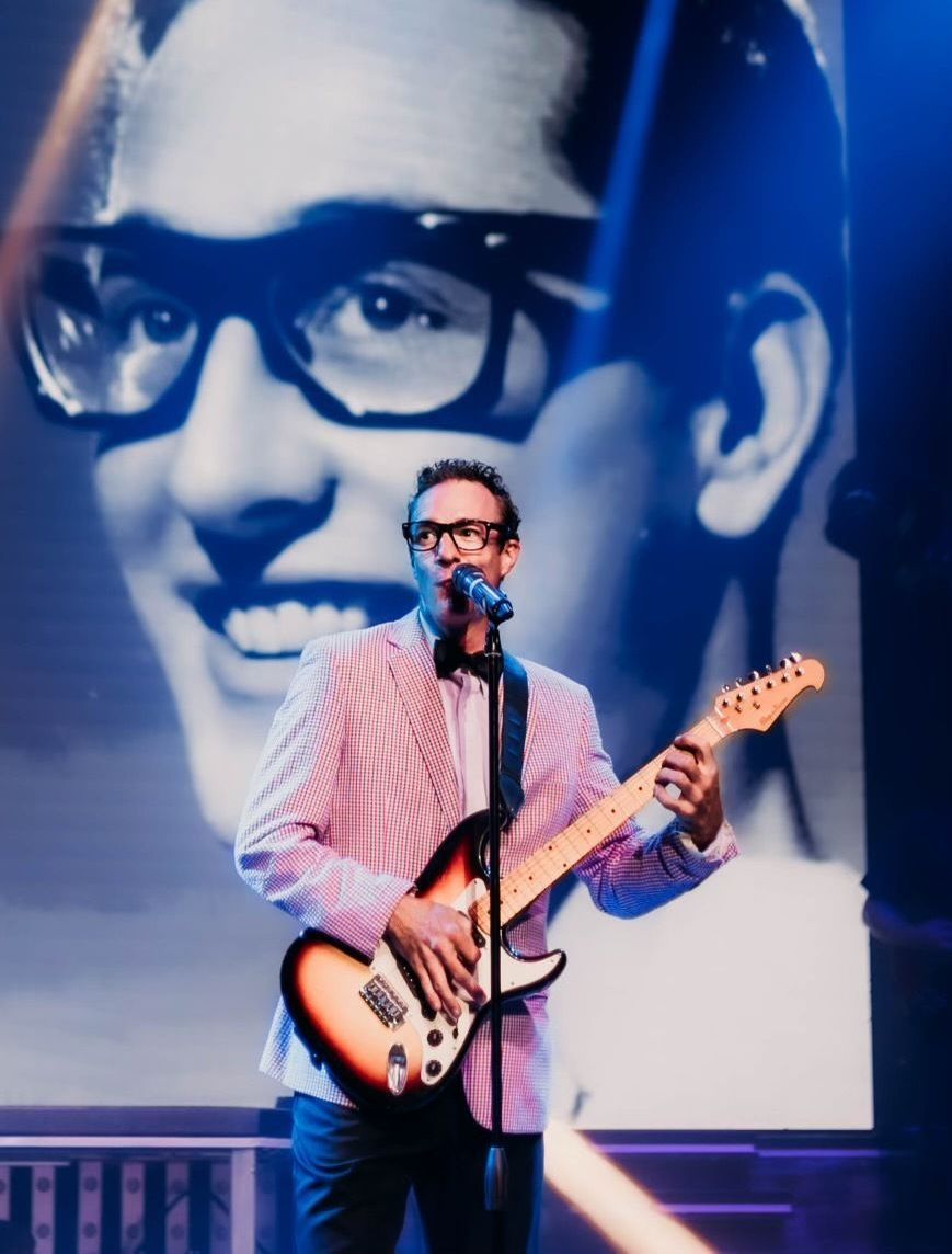 A performer playing an electric guitar onstage with a large black-and-white projected image of Buddy Holly behind them.