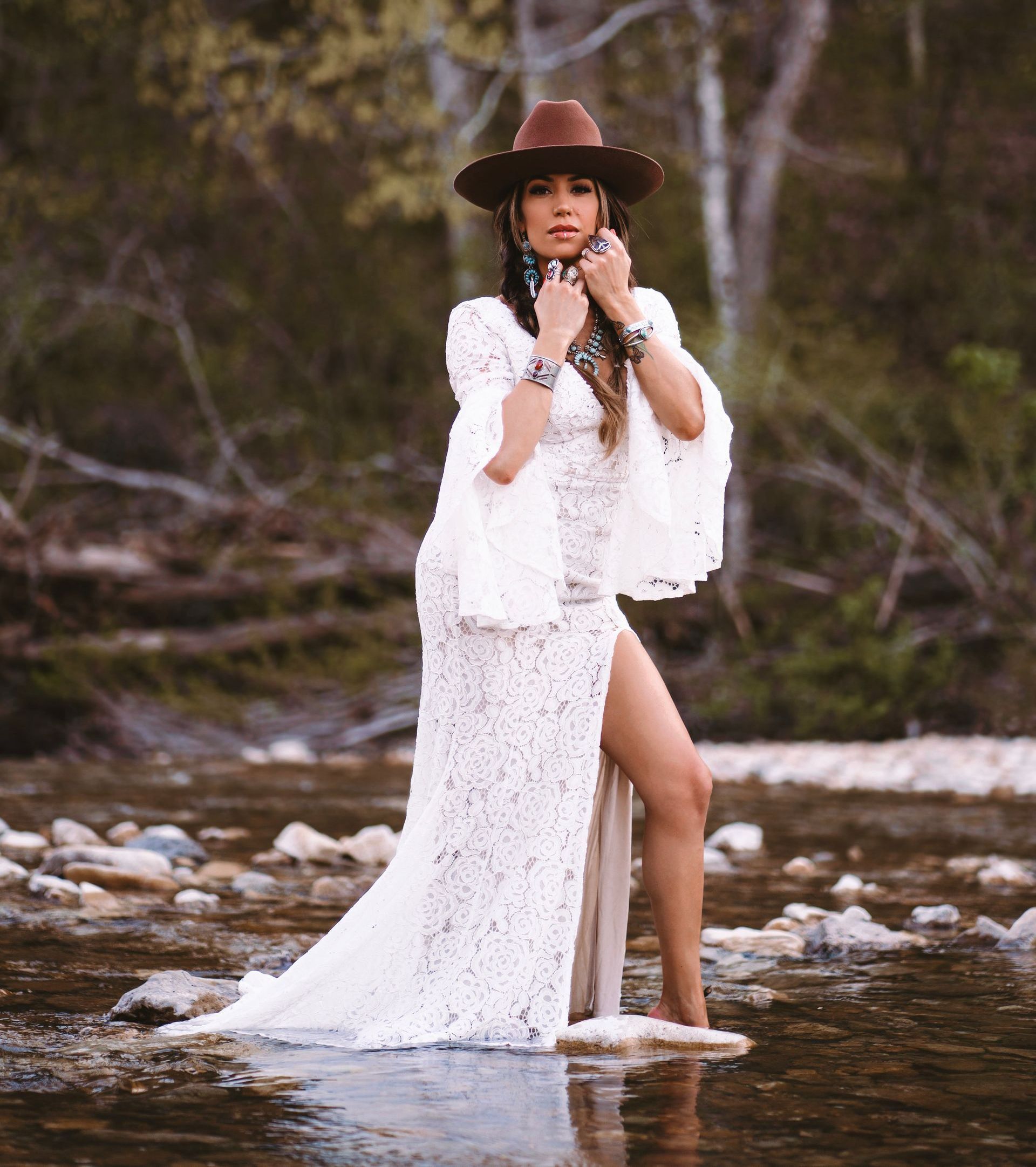 A person wearing a white lace dress and brown cowboy hat stands in a shallow stream, hands near their face.
