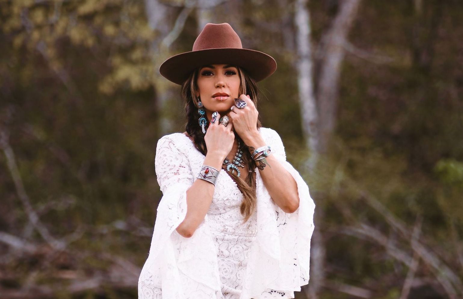 A person wearing a brown cowboy hat, white lace top, and turquoise jewelry, posing in a wooded outdoor setting.