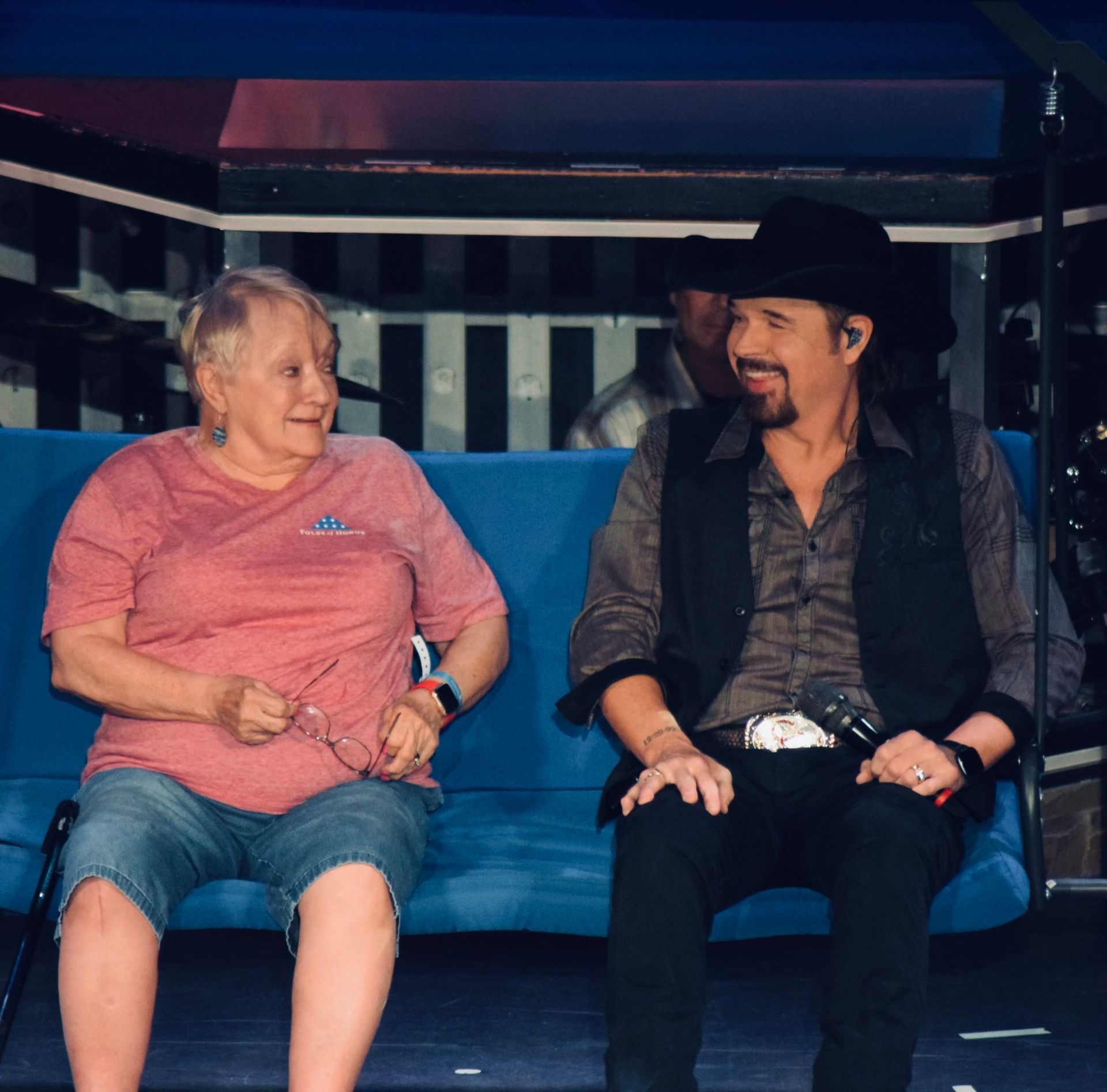 A person in a pink shirt and a person in a black hat and vest sit together on a blue bench, talking and smiling.