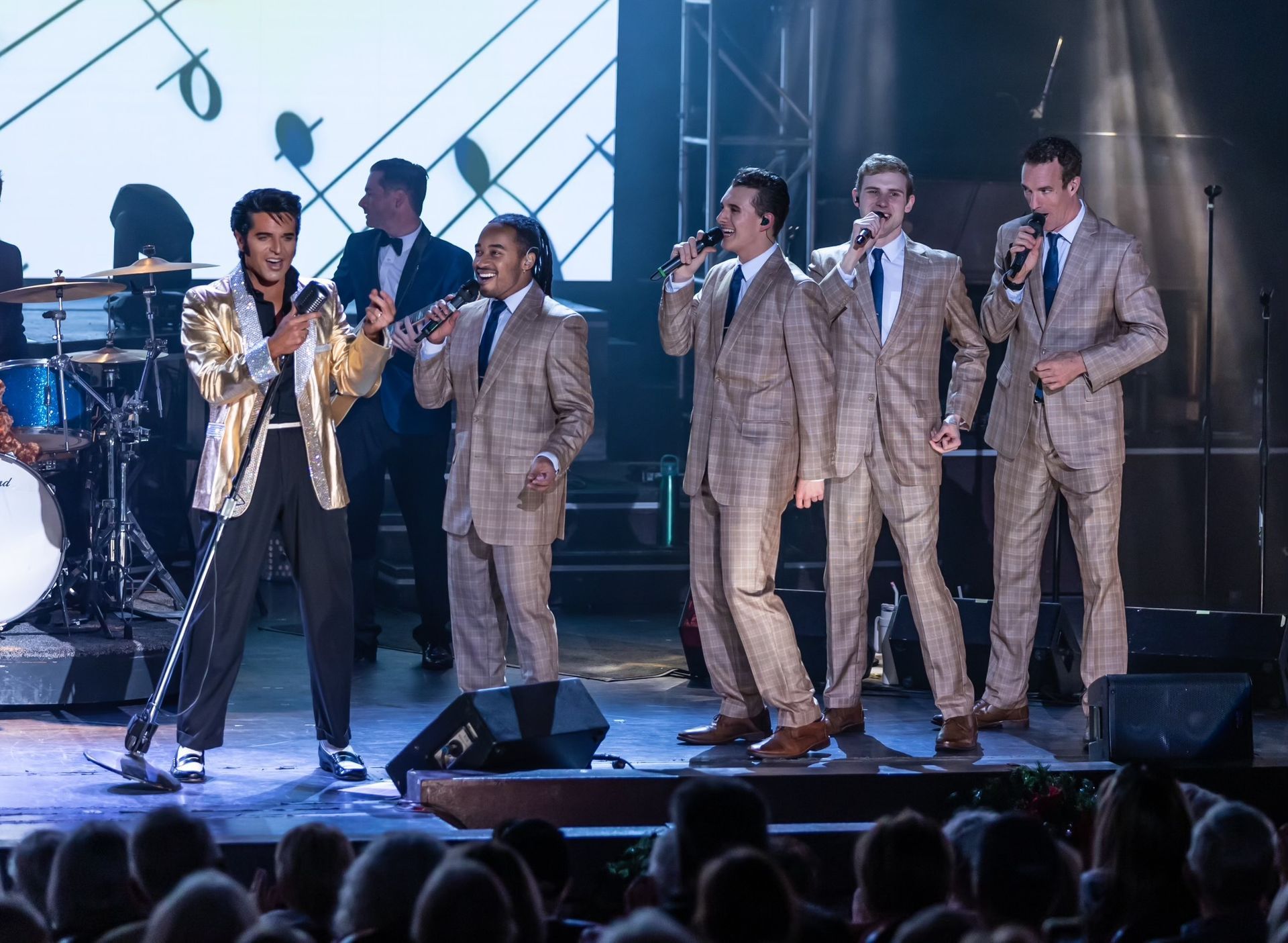 Five singers on a dimly lit stage perform with microphones, four wearing matching tan suits and one in a gold jacket.
