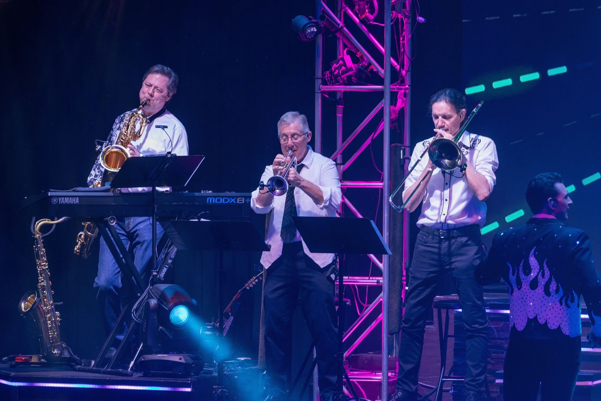 Three musicians play a saxophone, trumpet, and trombone on a stage with a metal truss, under bright stage lighting.
