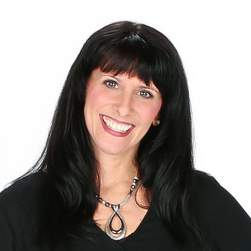 Smiling woman with long, dark hair wearing a black top and a teardrop pendant necklace, set against a white background.