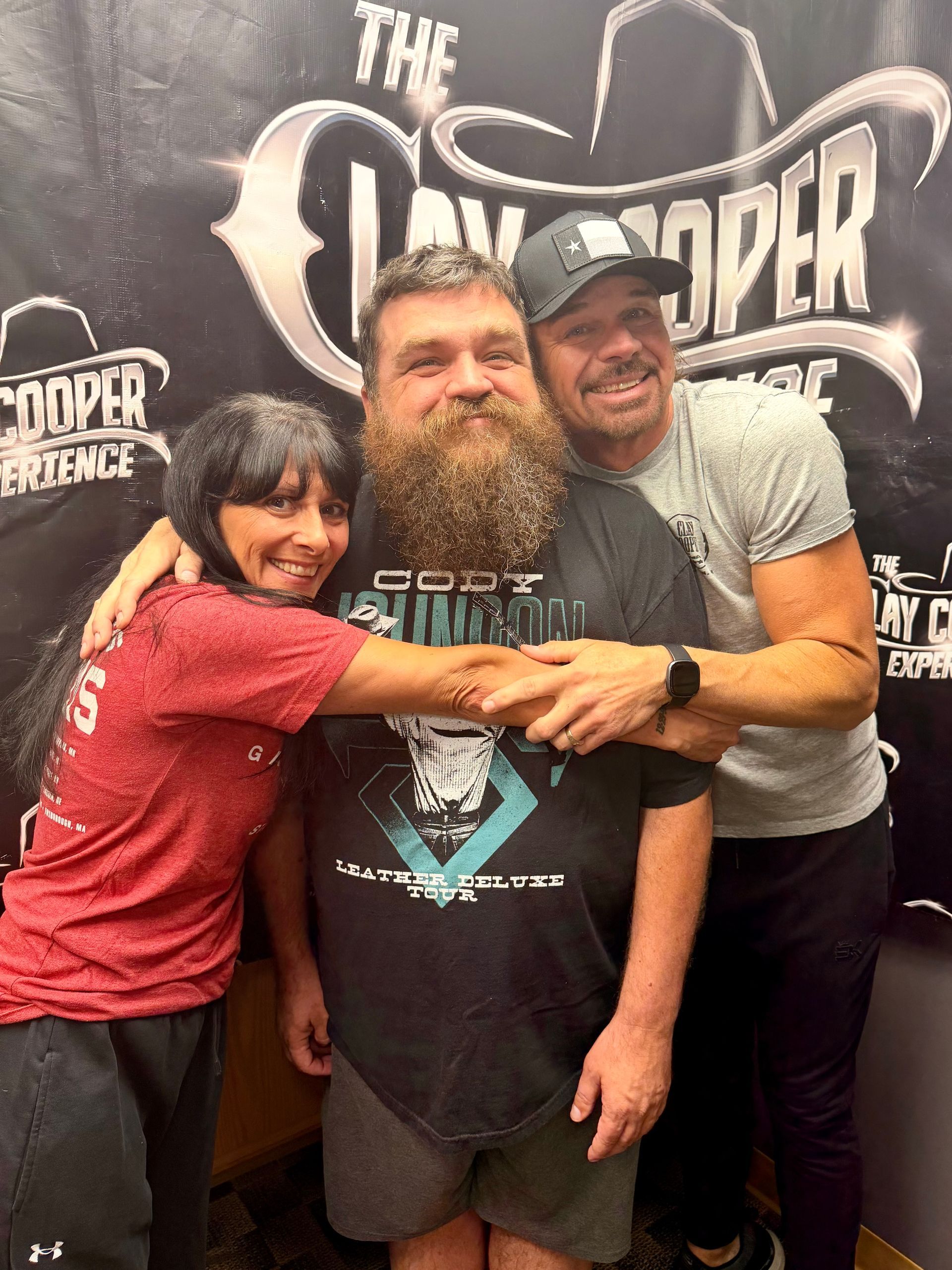 A woman with black hair is hugging a man with a beard who is also being hugged on the other side by a man in a baseball hat