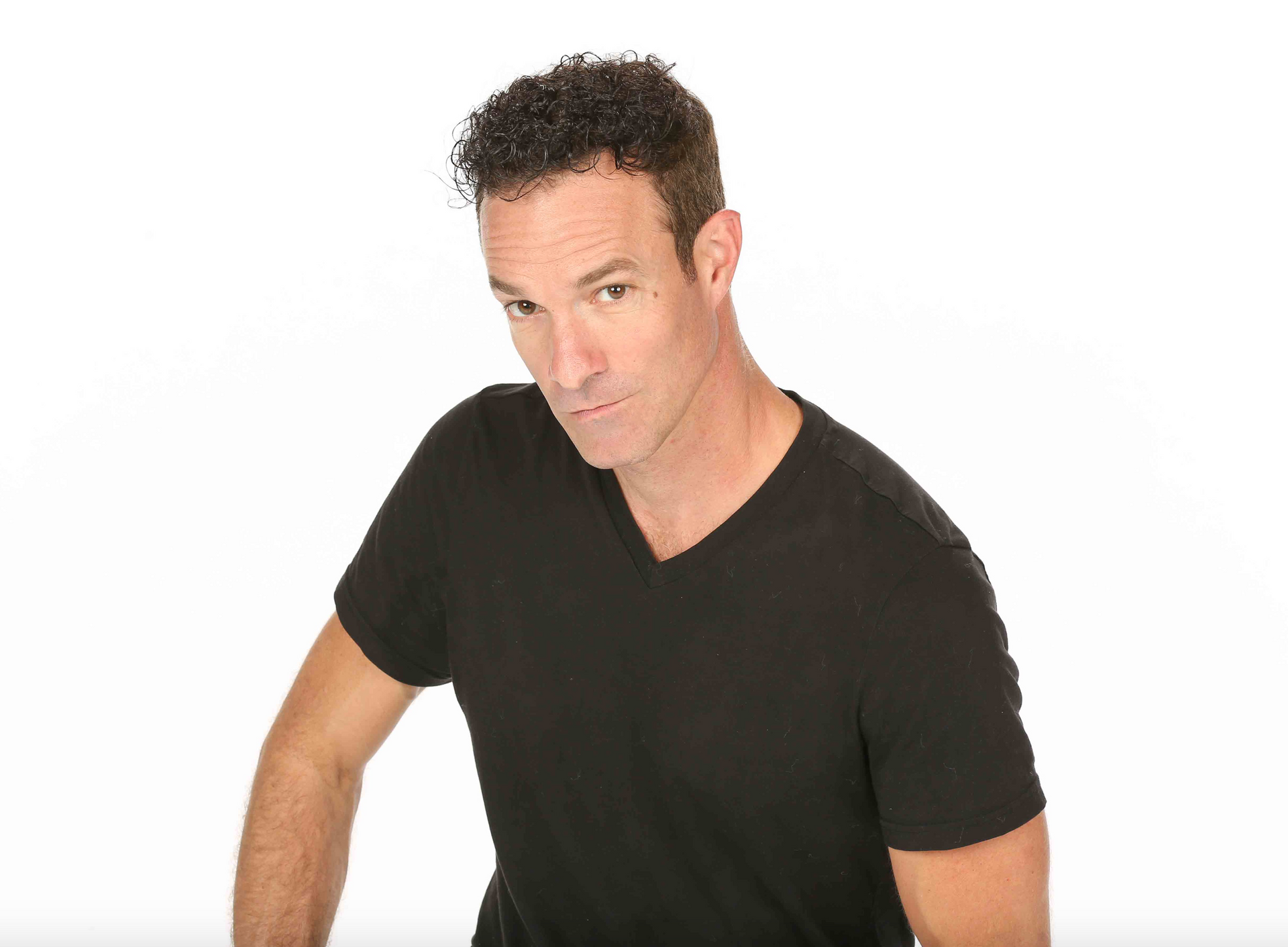 A person with short, dark curly hair wearing a black V-neck shirt, posing against a white background.