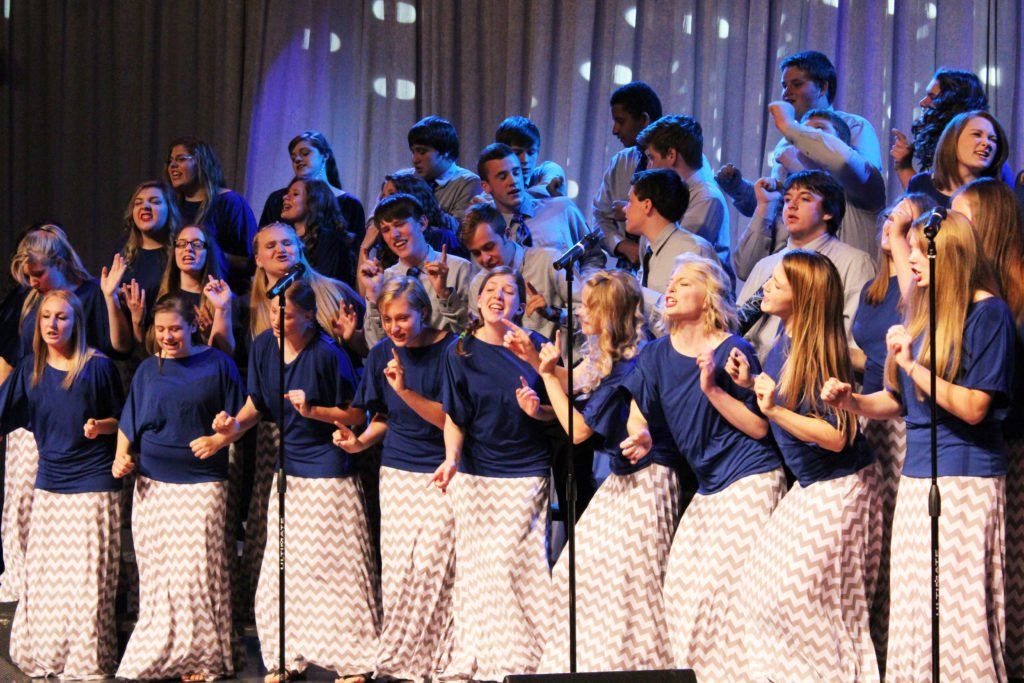 A choir wearing blue shirts and patterned skirts performing on a stage with a curtain backdrop and bright lights.