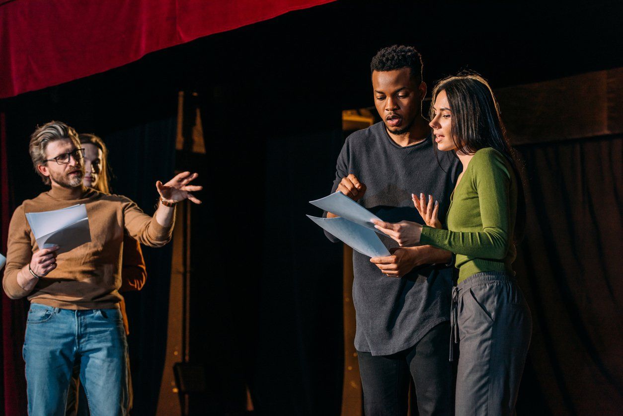 Three people on a theater stage read from scripts, with one person gesturing while speaking to the others.