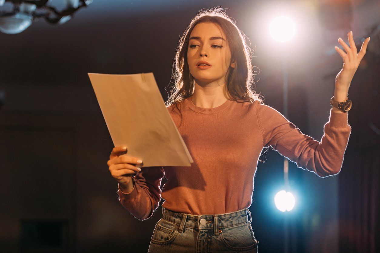 A person in a pink shirt holds a script, one hand raised as if acting on a stage with dramatic lighting.