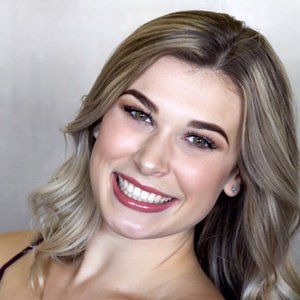 A headshot of a smiling person with wavy, highlighted blonde hair against a neutral gray background.