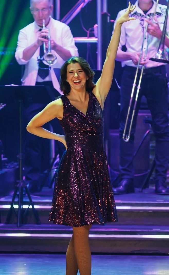 A smiling person in a dark sequined dress poses with one arm raised on a stage in front of a live band.