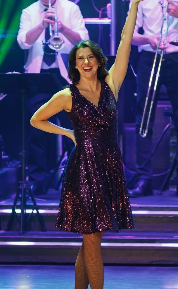 A smiling performer in a sparkly purple dress stands on a stage with an arm raised, backed by a band with brass instruments.