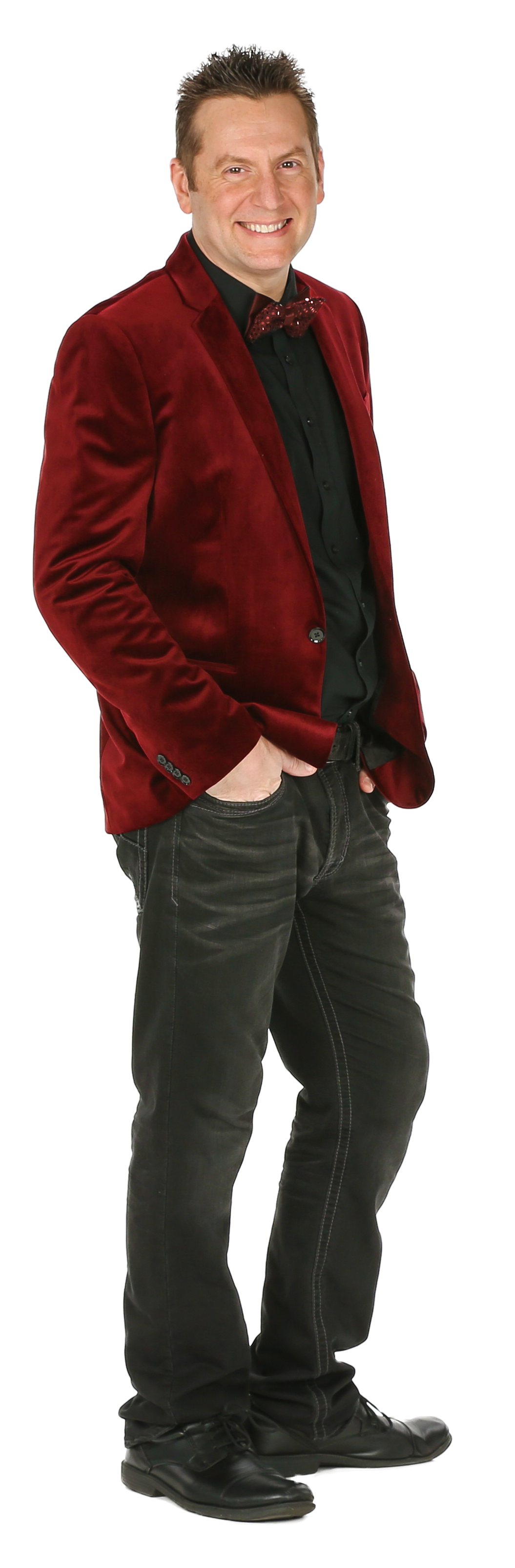 A smiling person standing with hands in pockets, wearing a red velvet blazer, black shirt, and dark jeans.
