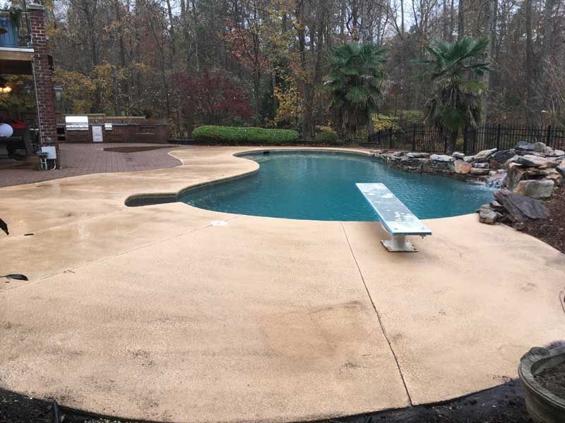 Backyard swimming pool with a diving board, tan deck, and trees in the background