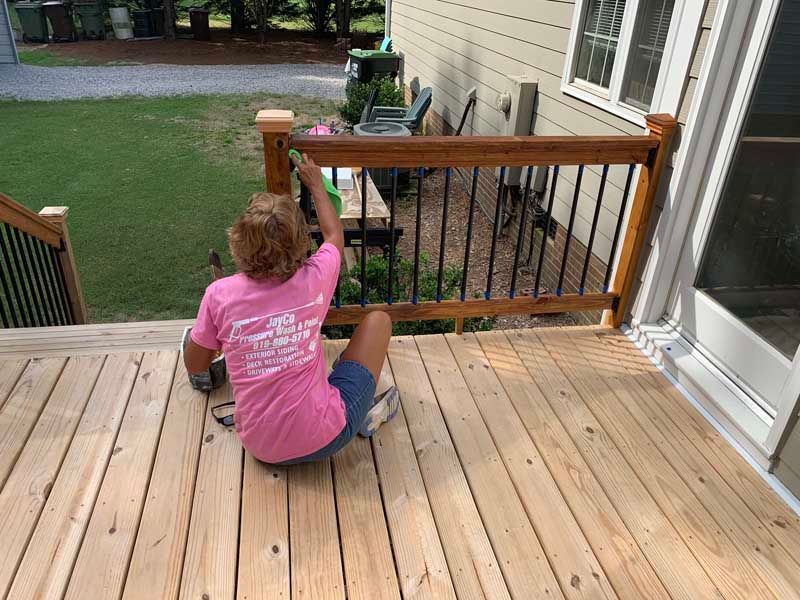 Person sitting on a wooden deck, painting the railing near a patio door.
