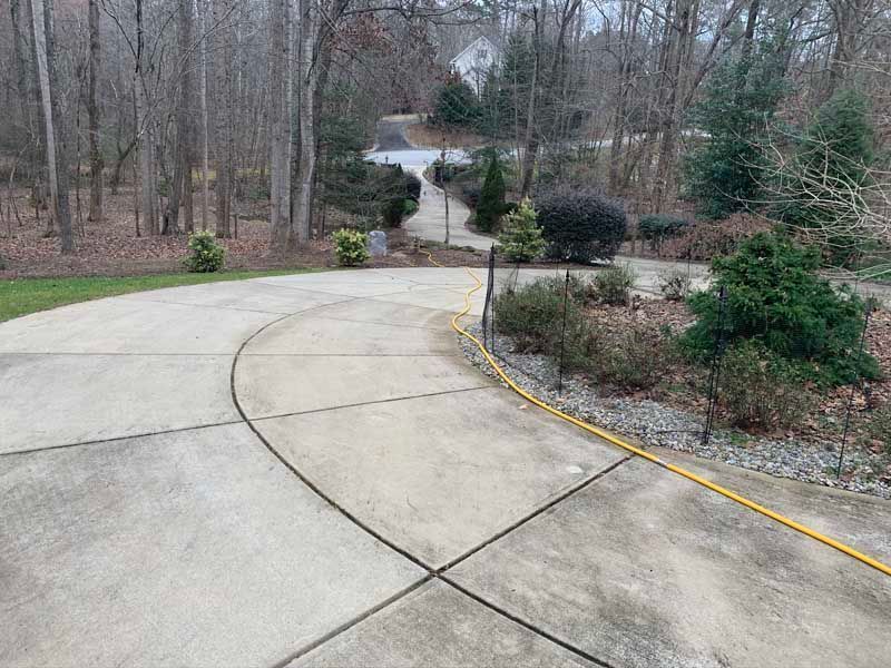 Curved concrete driveway leading to a wooded yard with shrubs and a yellow hose line along the edge.
