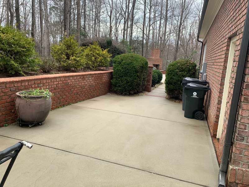 Brick patio walkway beside a house, with potted plants, shrubs, and a trash bin along a brick wall.