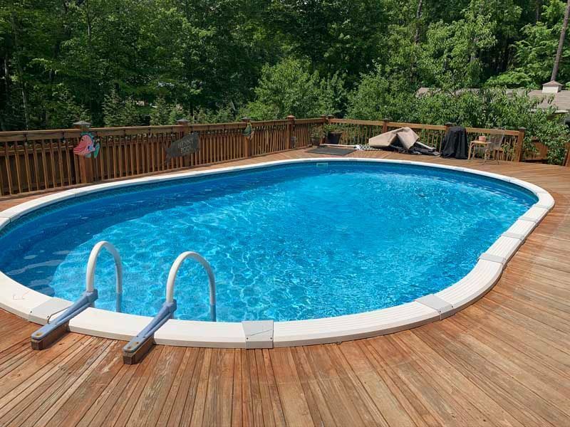 Blue swimming pool on a wooden deck with white coping, surrounded by trees and railing.