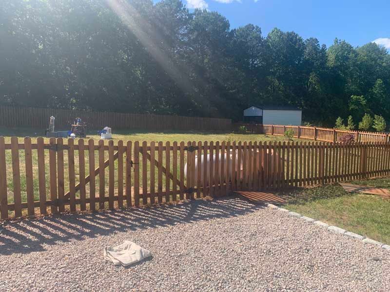 Wooden fence around a grassy field with people in the distance and trees beyond under bright sun.