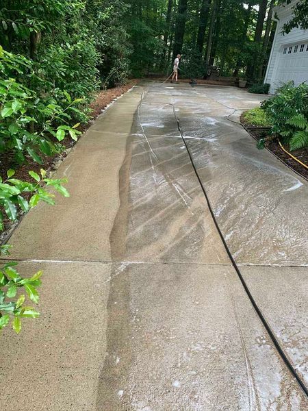 Wet concrete driveway beside lush green trees and a white house, with a person in the distance.