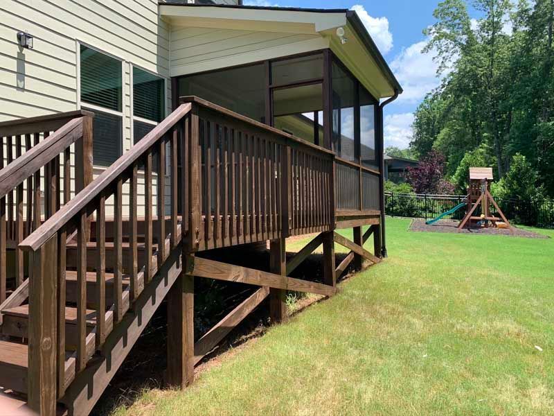 Wooden porch with stairs on a house beside a grassy yard and a playground swing set in the background.