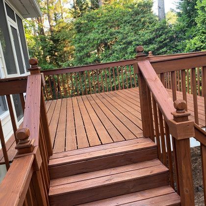 Wooden deck stairs leading to a spacious brown deck surrounded by lush green trees.