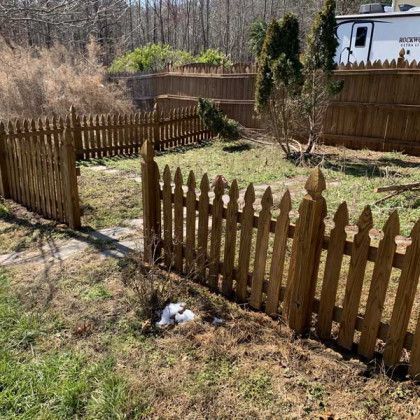 Small fenced backyard with brown picket fencing, patchy grass, shrubs, and a white RV in the background