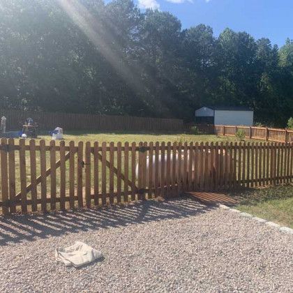 Wooden fence along a gravel path with a small building and trees in bright sunlight