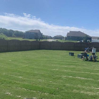 Sunny backyard with mowed lawn, wooden fence, and patio chairs under a blue sky.