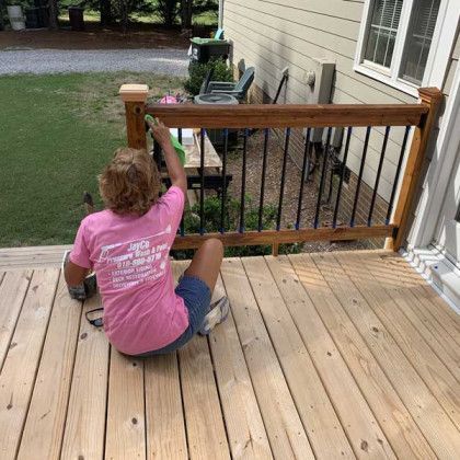 Person in a pink shirt kneels on a wooden deck, working on a railing beside a house.