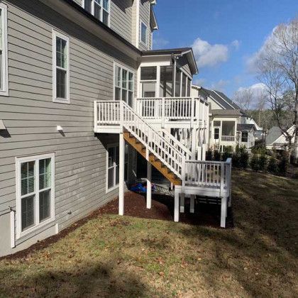 Backyard view of a gray house with a white deck, stairs, and railing above a grassy yard.