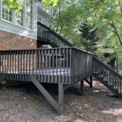 Raised wooden deck with black railings and stairs beside a brick house in a leafy yard