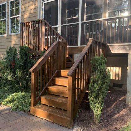 Wooden exterior stairs leading to a raised porch beside a house, with shrubs on both sides.